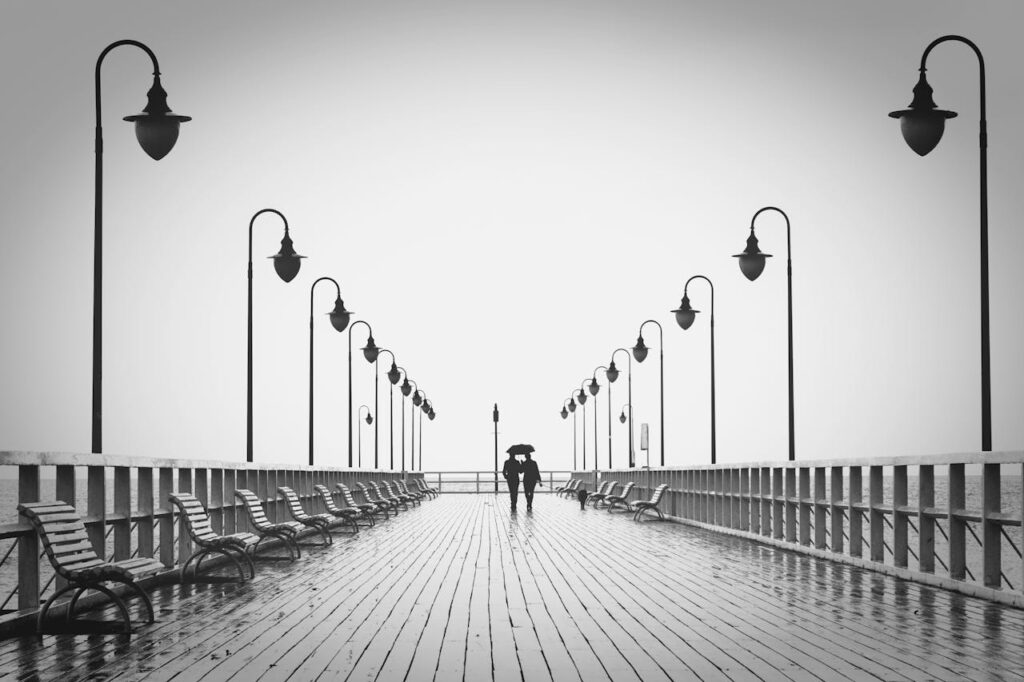 A couple holding umbrellas walks on a rainy boardwalk, embodying romance and tranquility.
